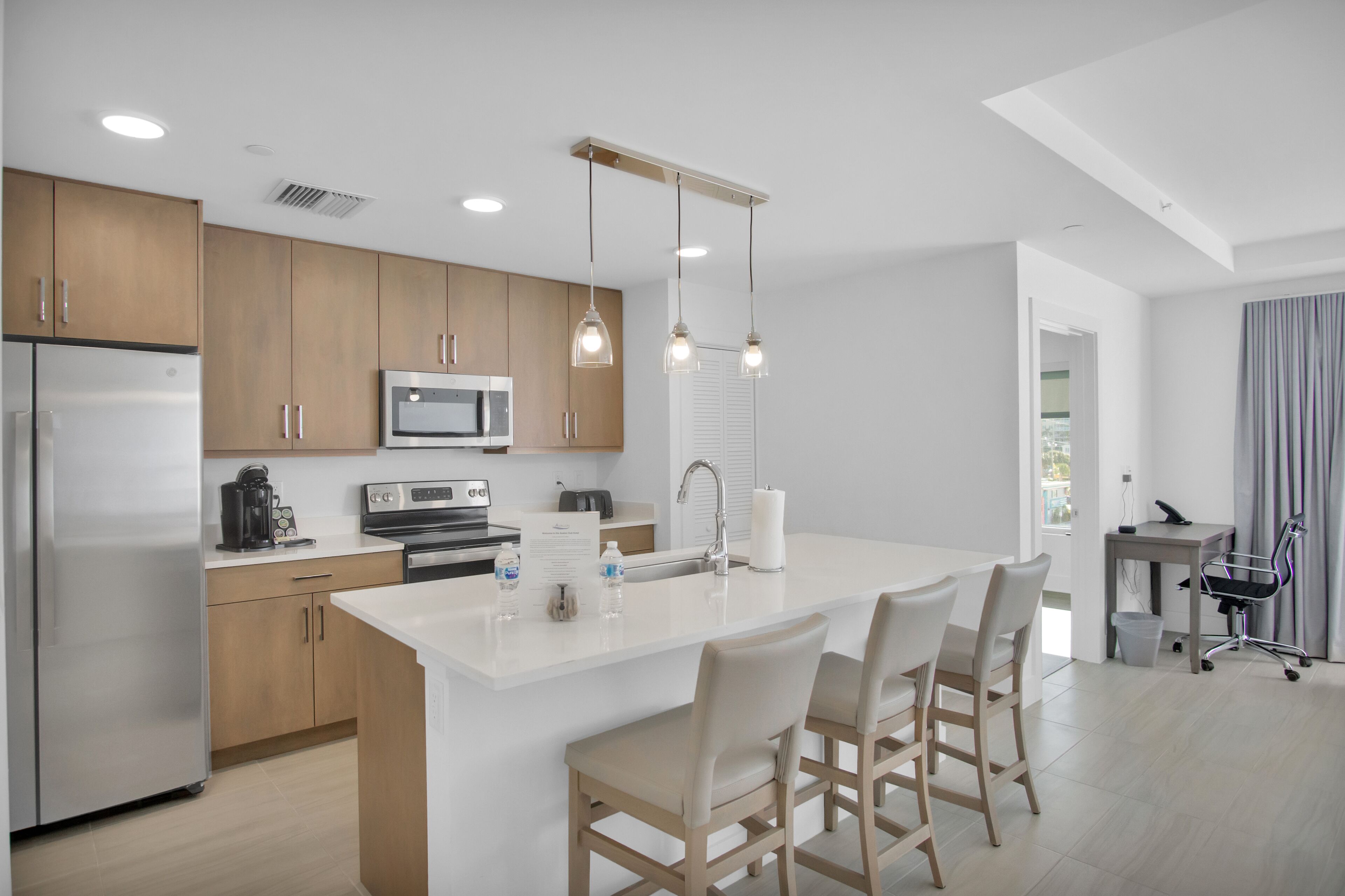 A modern kitchen with wooden cabinets, stainless steel appliances, a large island with bar stools, pendant lighting, and a view to the living area.