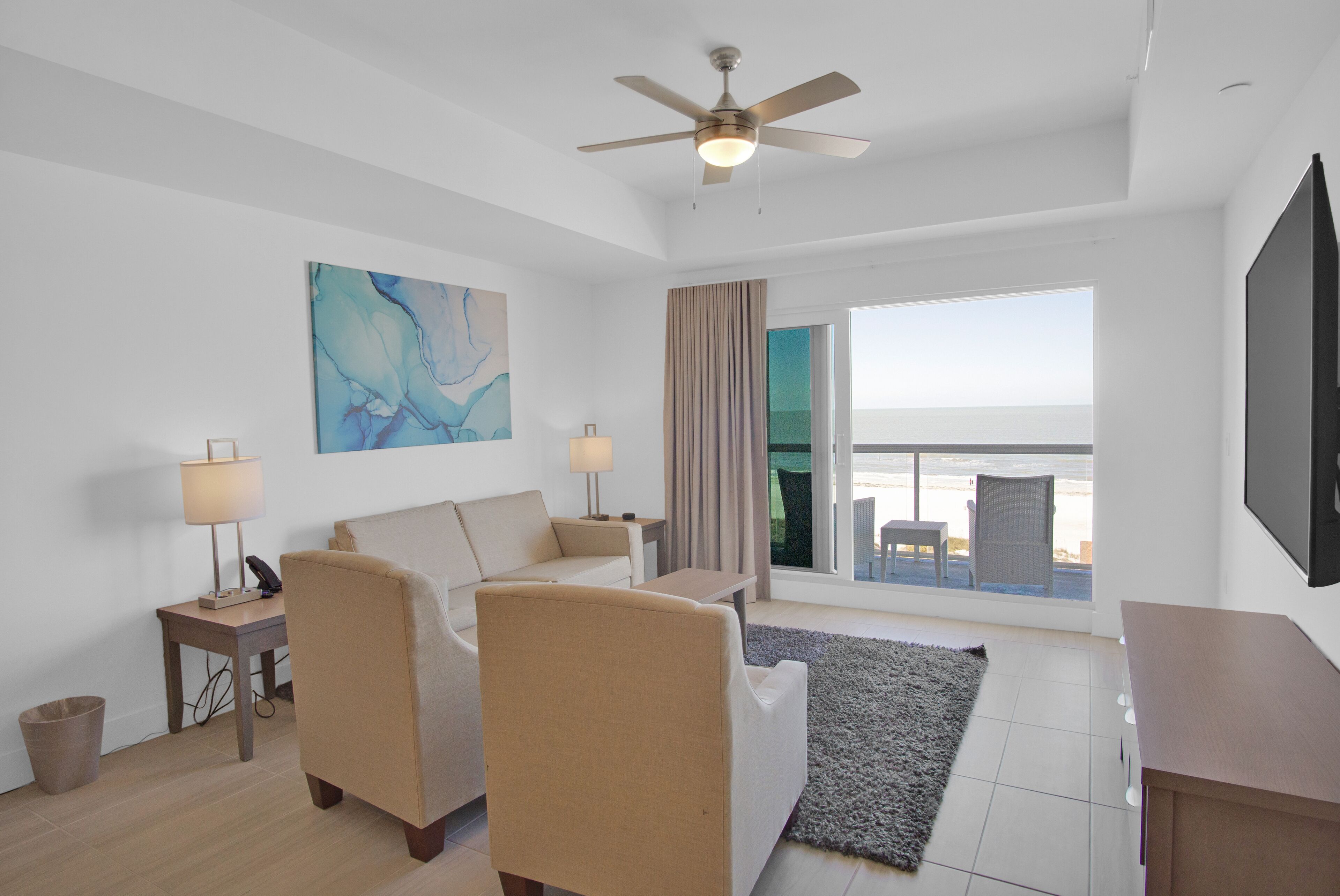A modern hotel room with a large window, a ceiling fan, a flat-screen TV, a sofa, a coffee table, and a painting on the wall.
