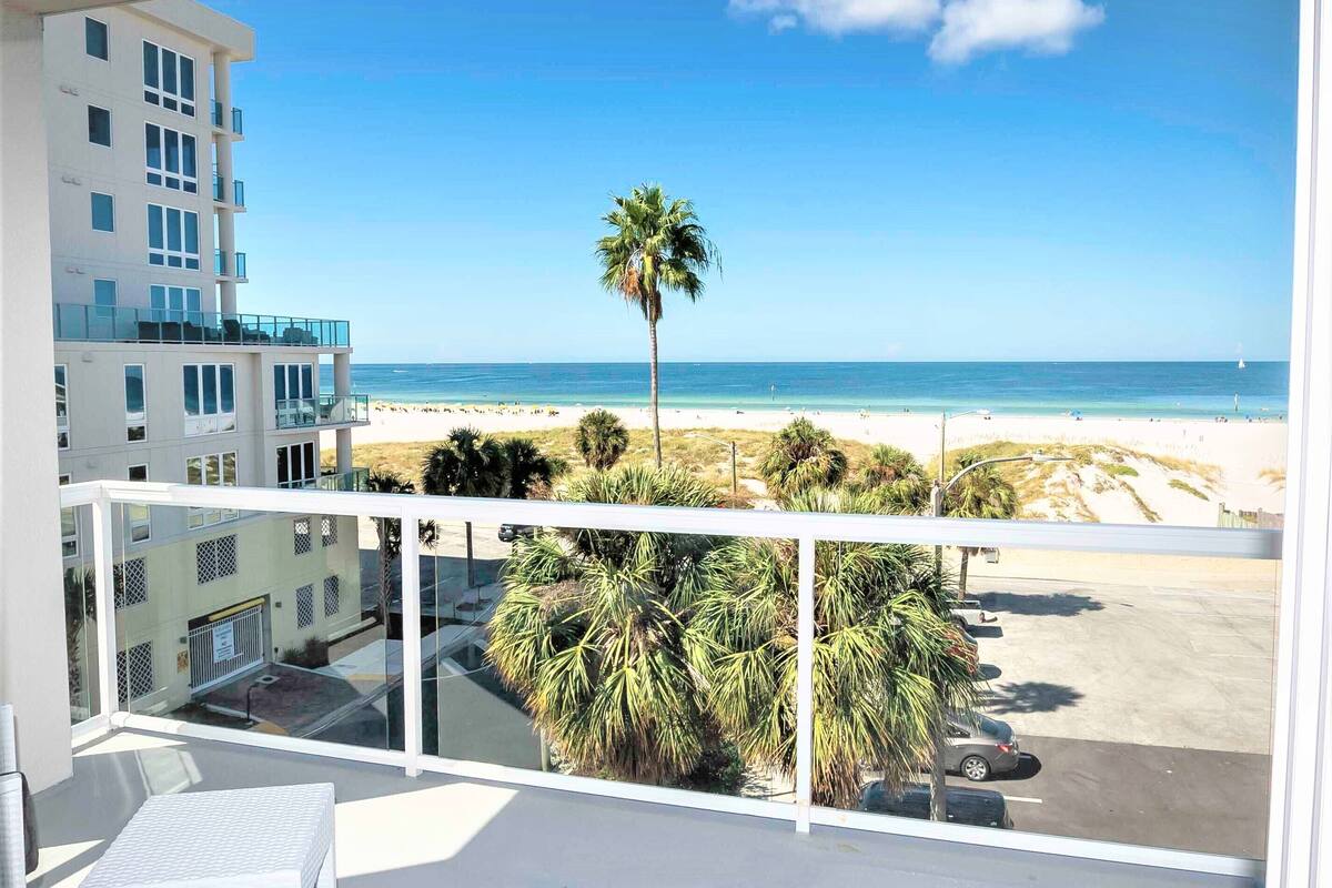 A balcony with a view of a beach, palm trees, and a building with multiple windows.
