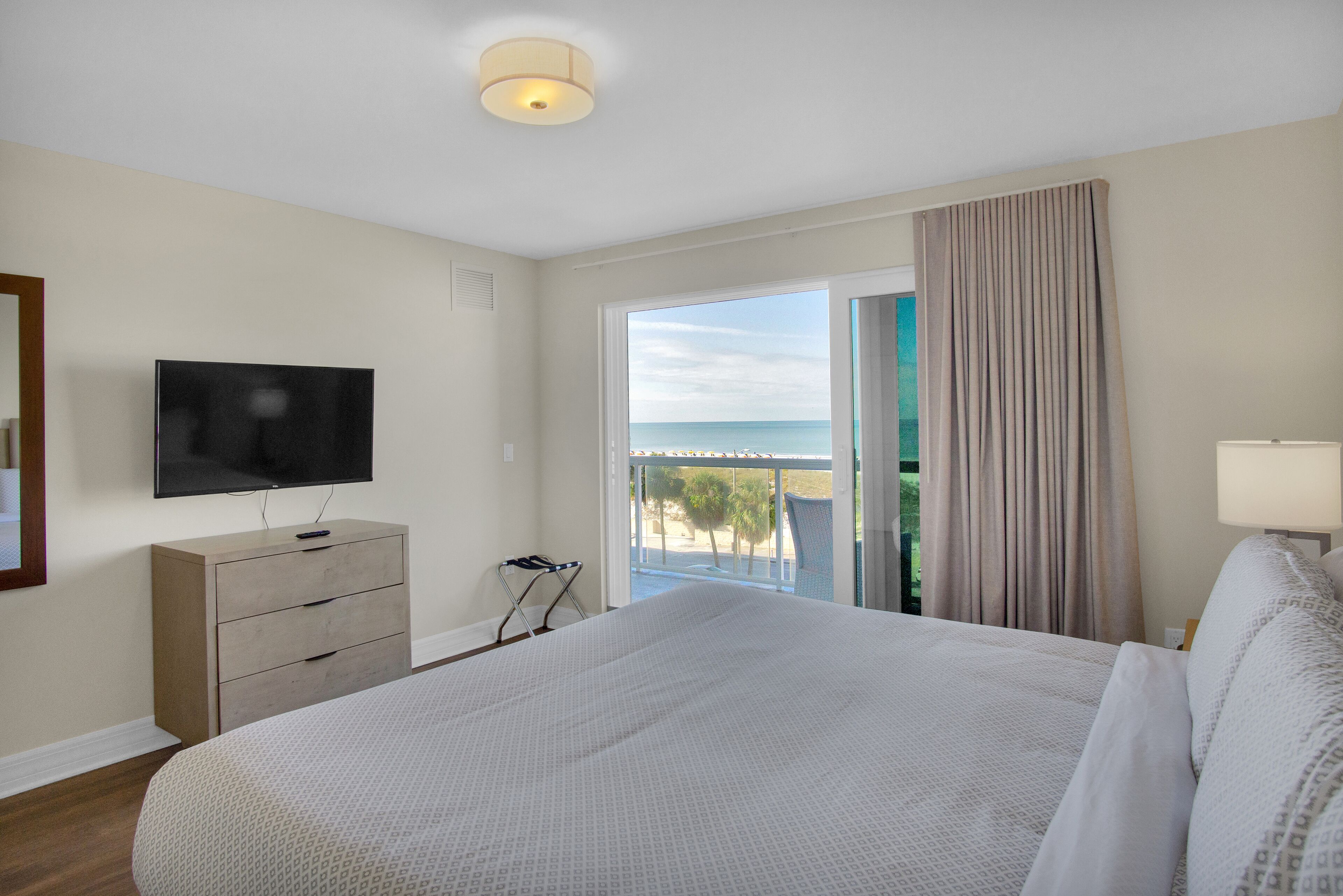 Habitación de hotel con una cama grande, un televisor en una cómoda, un balcón con vistas al mar y una ventana con cortinas.