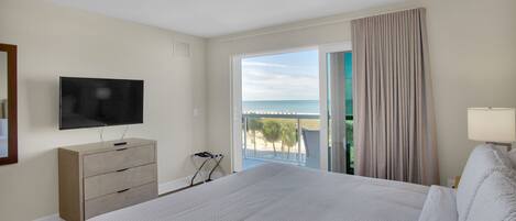 Habitación de hotel con una cama grande, un televisor en una cómoda, un balcón con vistas al mar y una ventana con cortinas.