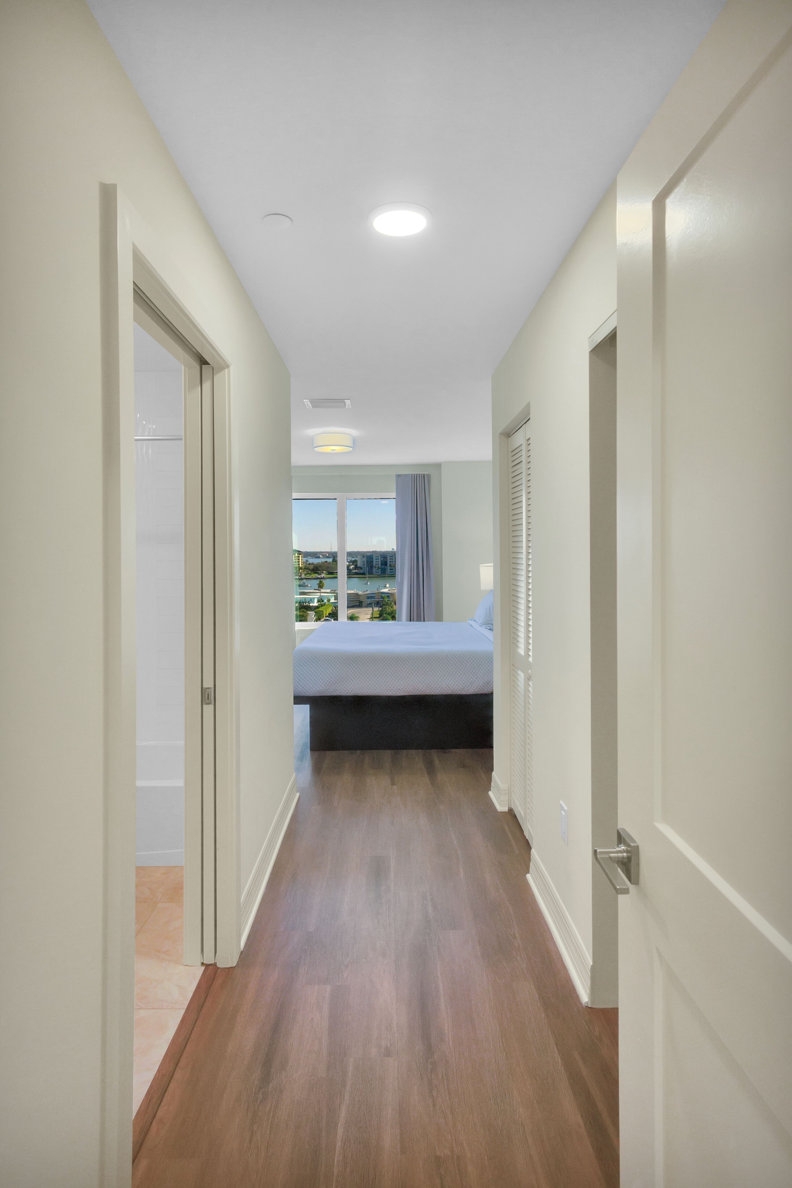 A hallway with wooden flooring leading to a bedroom with a bed and a window.