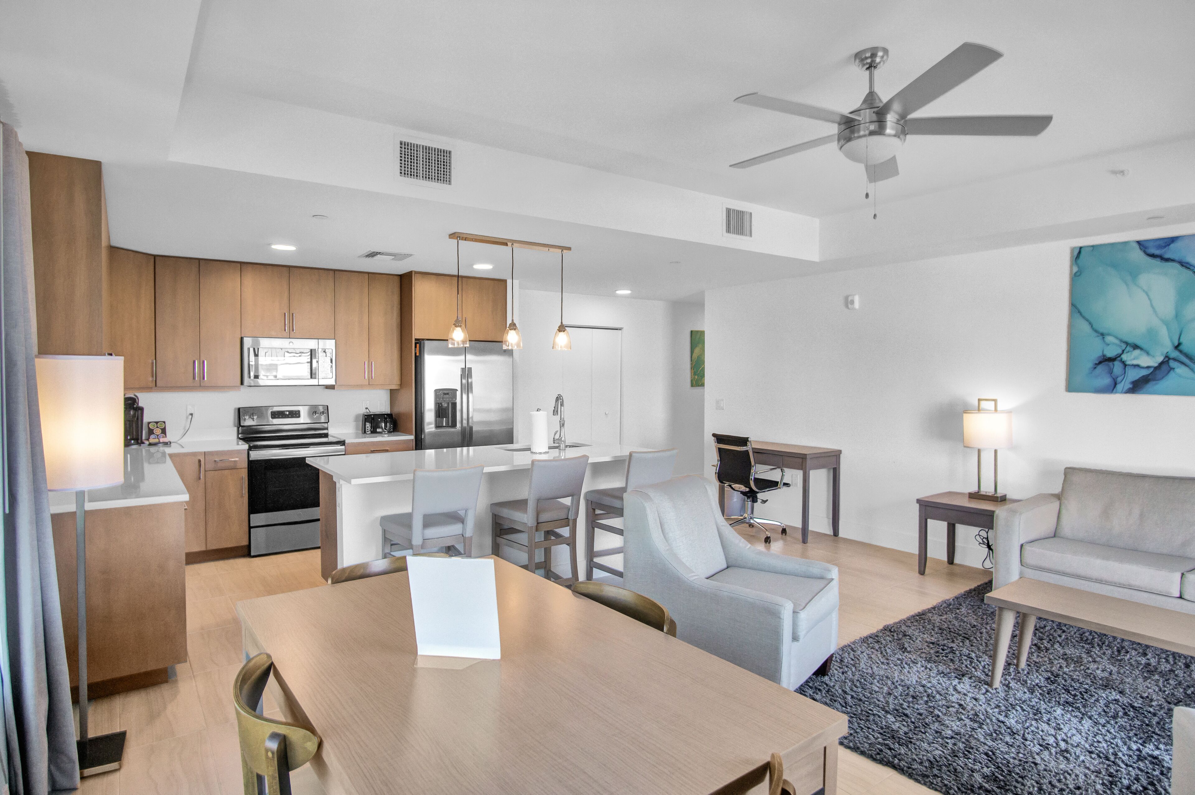 A modern kitchen with stainless steel appliances, a dining area with a table and chairs, a living room with a sofa, and a ceiling fan.