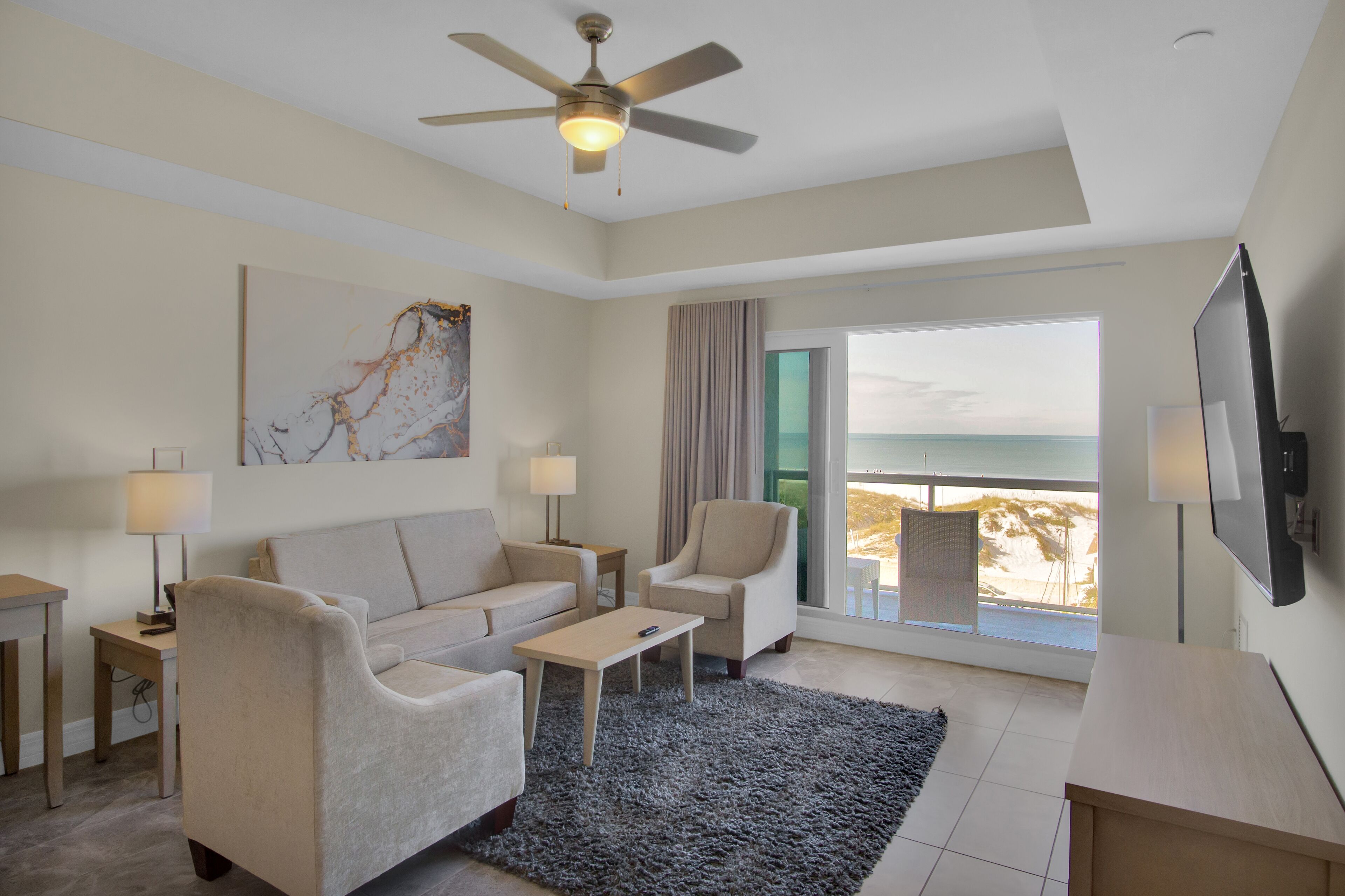 A modern hotel room with a sofa, armchair, coffee table, and a TV. There's a large window with a view of the beach and a ceiling fan.