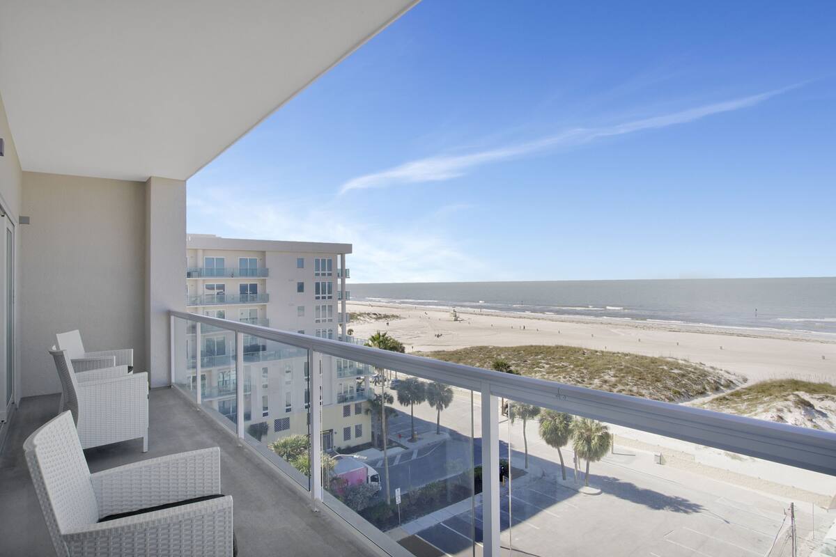 A balcony with white wicker furniture overlooking a beach and a parking area.