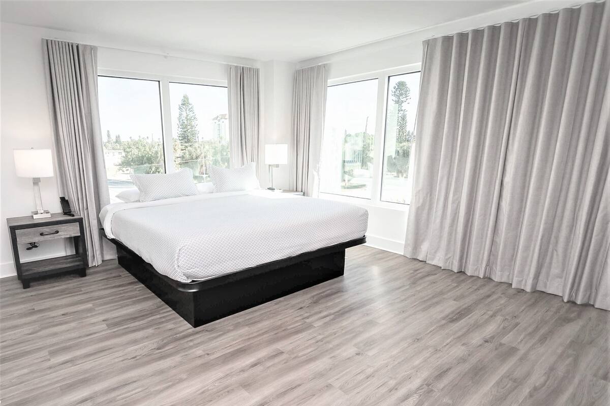 A modern bedroom with a large bed, two bedside tables, and a view of the outdoors through large windows.
