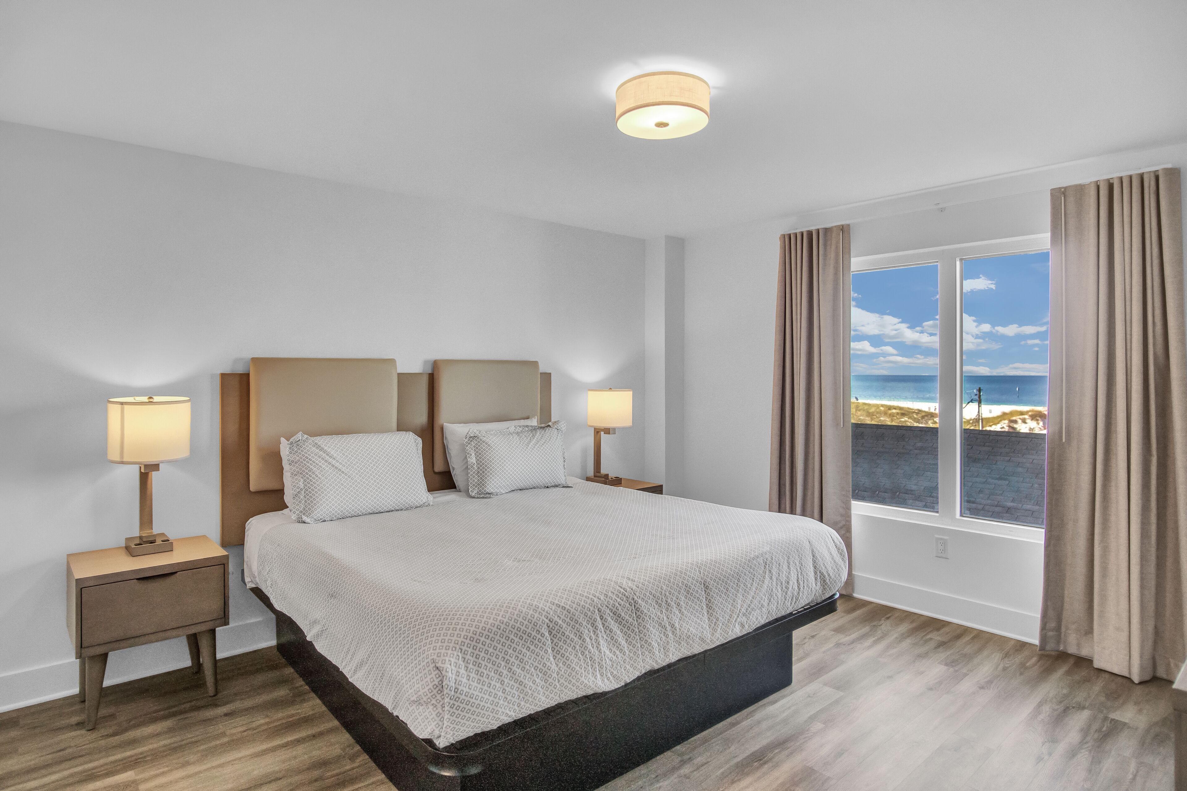 A bedroom with a large bed, two bedside tables with lamps, a window with curtains, and a view of the beach.