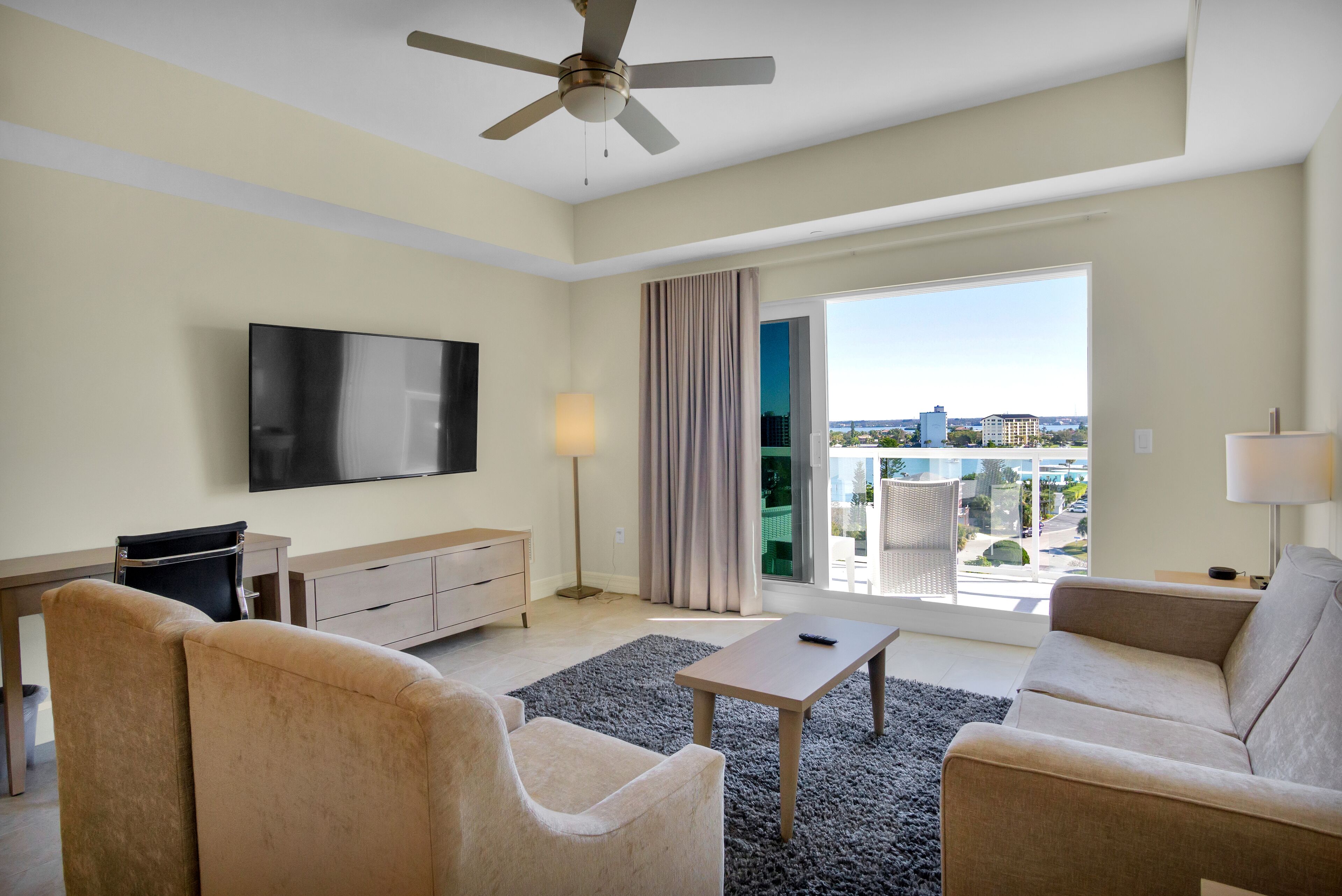 A modern hotel room with a flat-screen TV, two beige sofas, a coffee table, a ceiling fan, and a large window offering a city view.