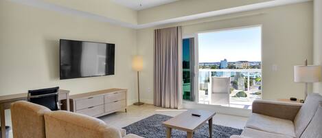 A modern hotel room with a flat-screen TV, two beige sofas, a coffee table, a ceiling fan, and a large window offering a city view.