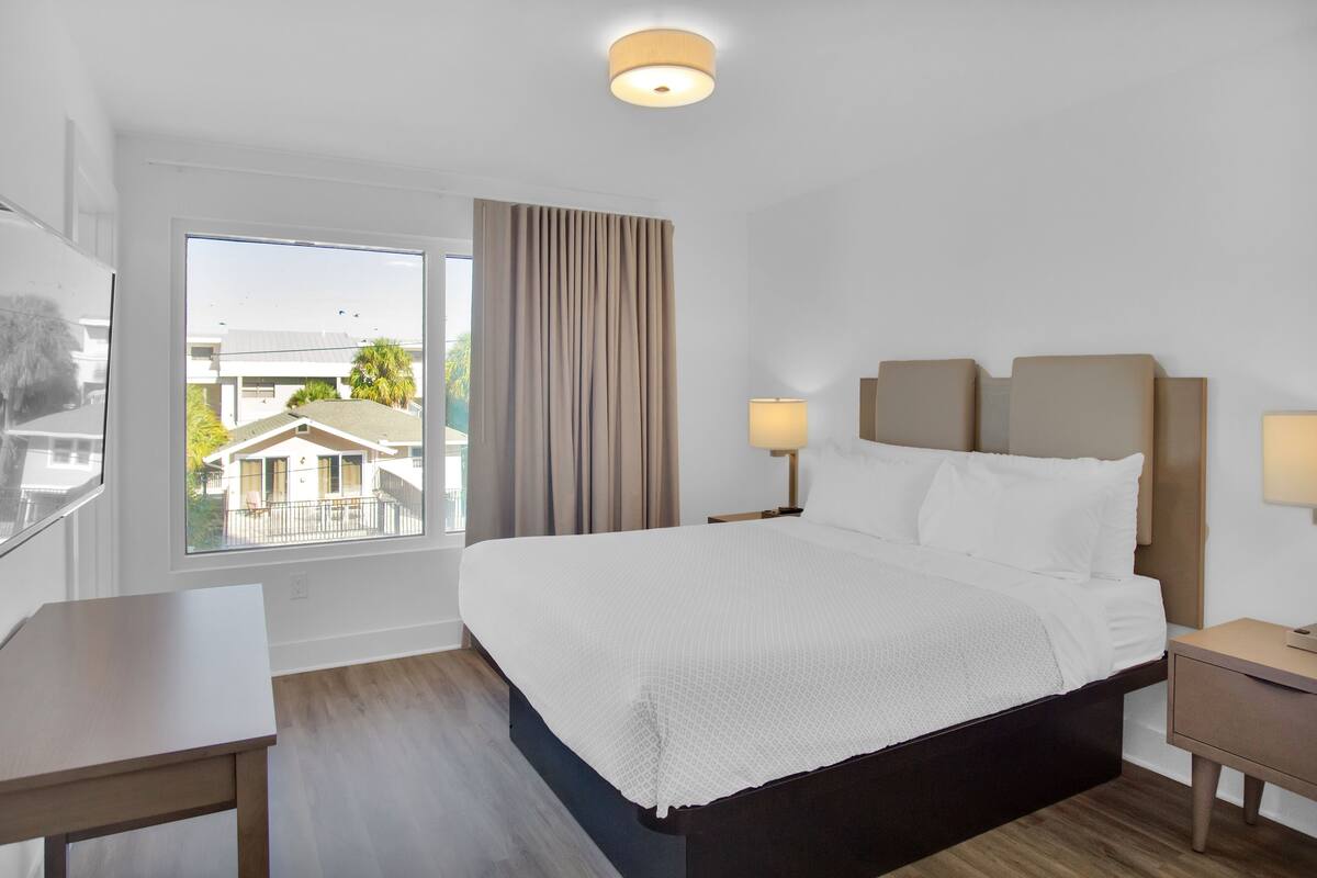 A modern hotel room with a large bed, bedside lamps, a small table, and a view of the outside.