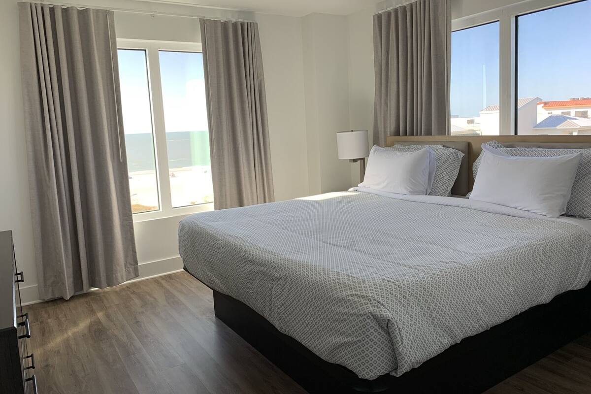 A hotel room with a large bed, a bedside table with a lamp, and a view of the sea through the windows.