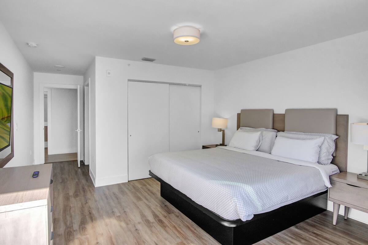 A bedroom with a bed, bedside tables, a nightstand, a lamp, a ceiling light, a door, and a wall-mounted artwork.
