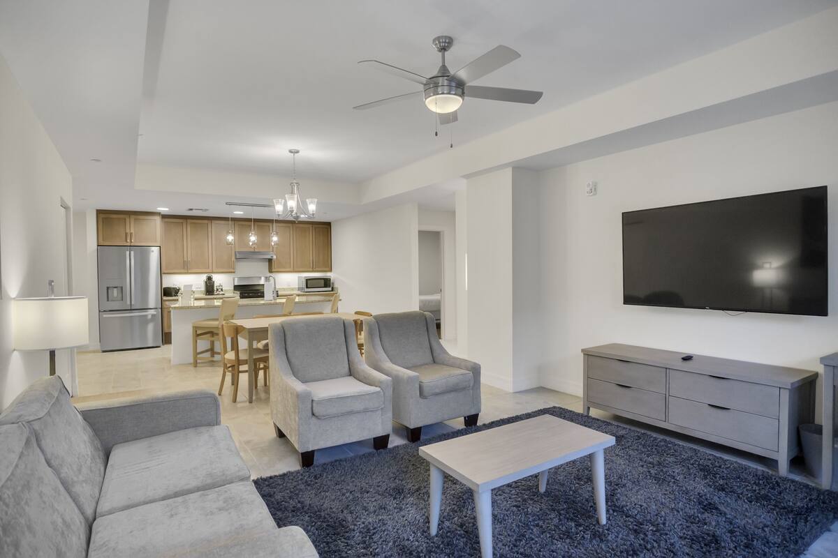 A modern living room with a grey sofa, a coffee table, and two armchairs. A flat-screen TV is mounted on the wall, and a ceiling fan is visible.