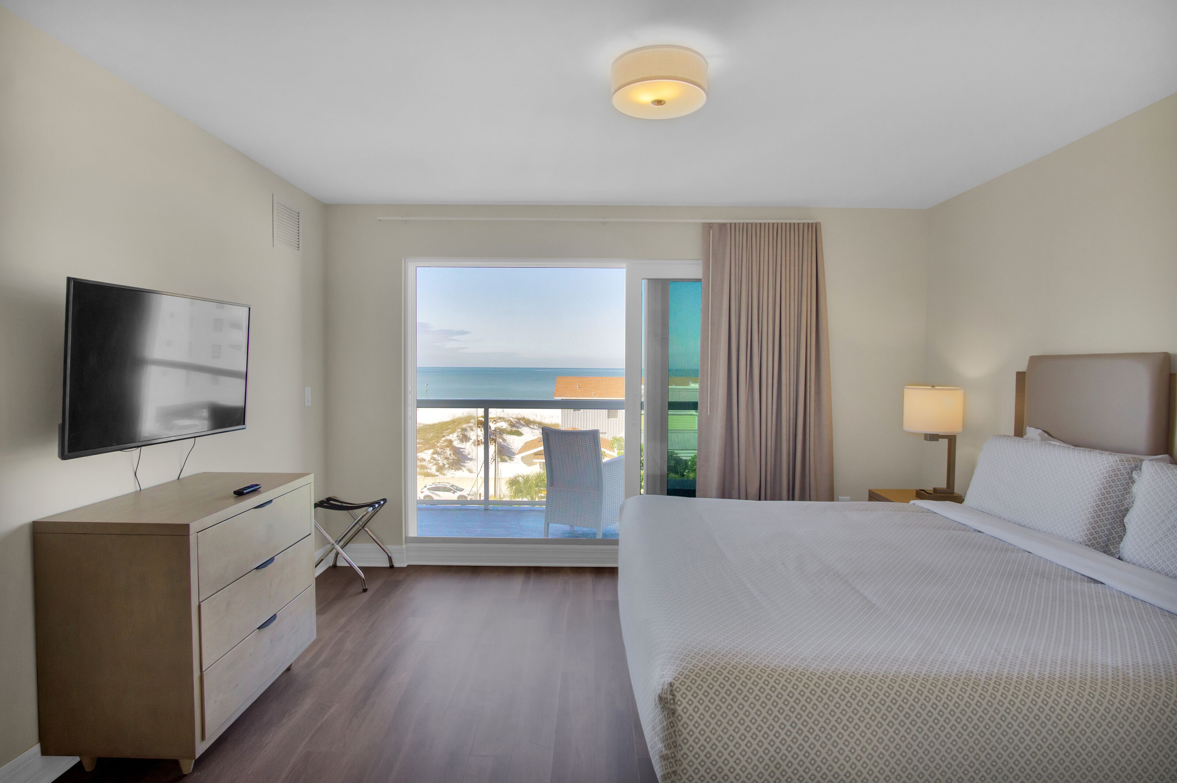 A hotel room with a bed, a TV, a dresser, a balcony with a view of the sea, and a ceiling light.