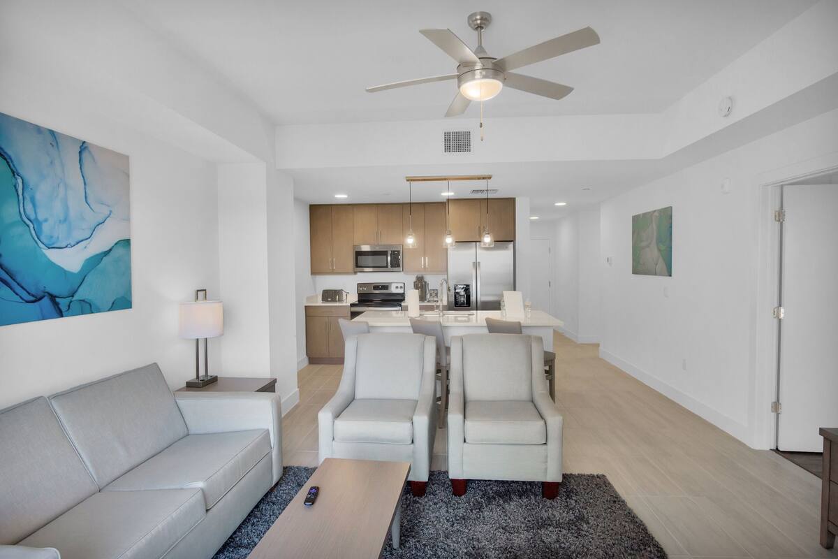 A modern living room with a sofa, a coffee table, and a dining area with chairs. There is a kitchen in the background with a microwave and a refrigerator.