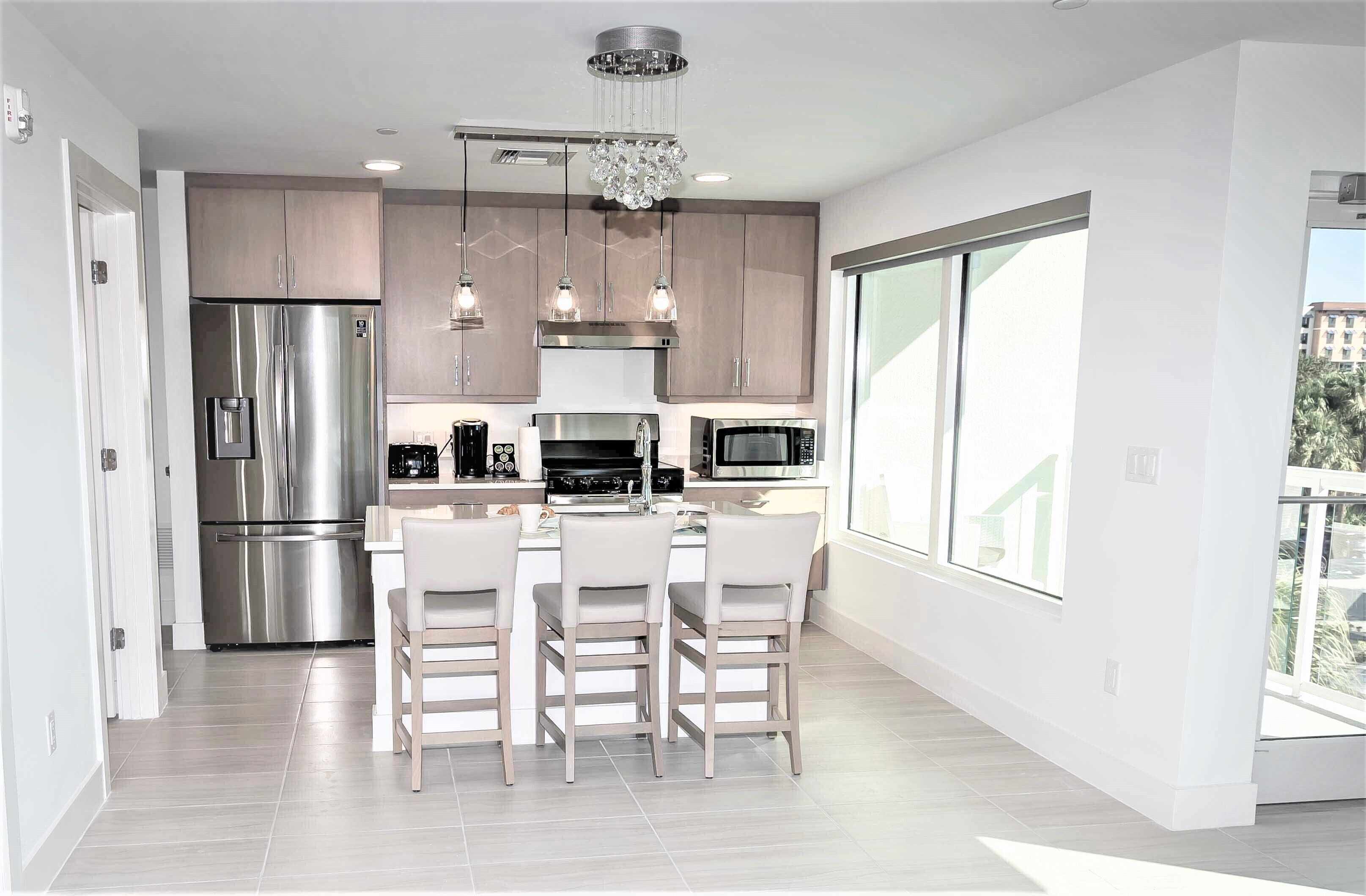 A modern kitchen with stainless steel appliances, a central island with bar stools, pendant lighting, and large windows offering a view.