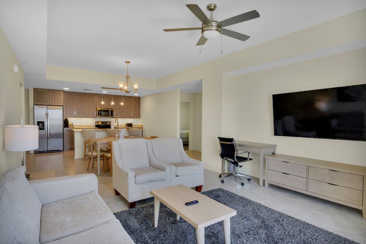 A modern living room with a sofa, coffee table, and a TV. There is a kitchen in the background with a dining area.