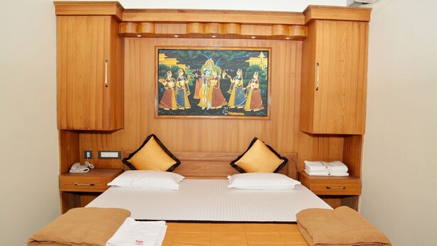 A hotel room with a large bed, two bedside tables, a wooden headboard with a painting, and matching wooden cabinets. - Anandha Grand (Tiruchendur)
