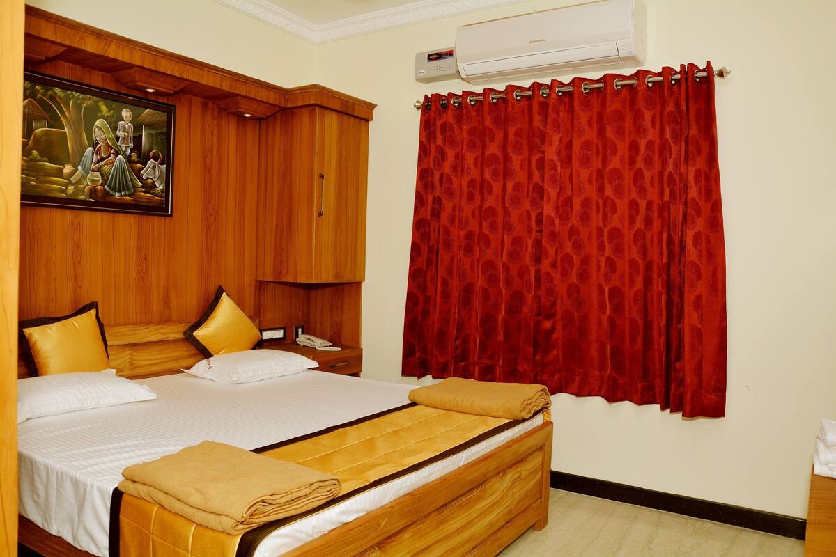 A hotel room with a bed, wooden wardrobe, red curtains, and a painting on the wall.