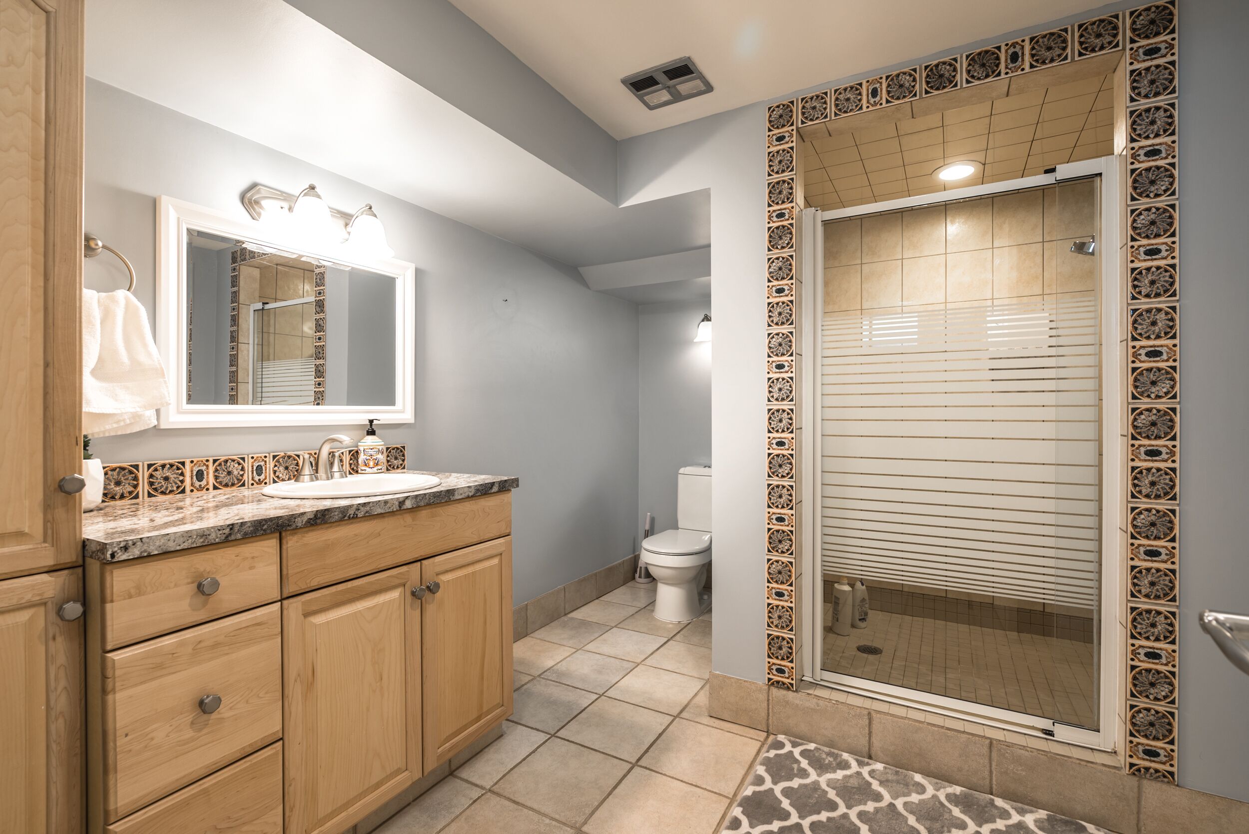 A bathroom with a sink, mirror, vanity, toilet, and shower with decorative tiles.