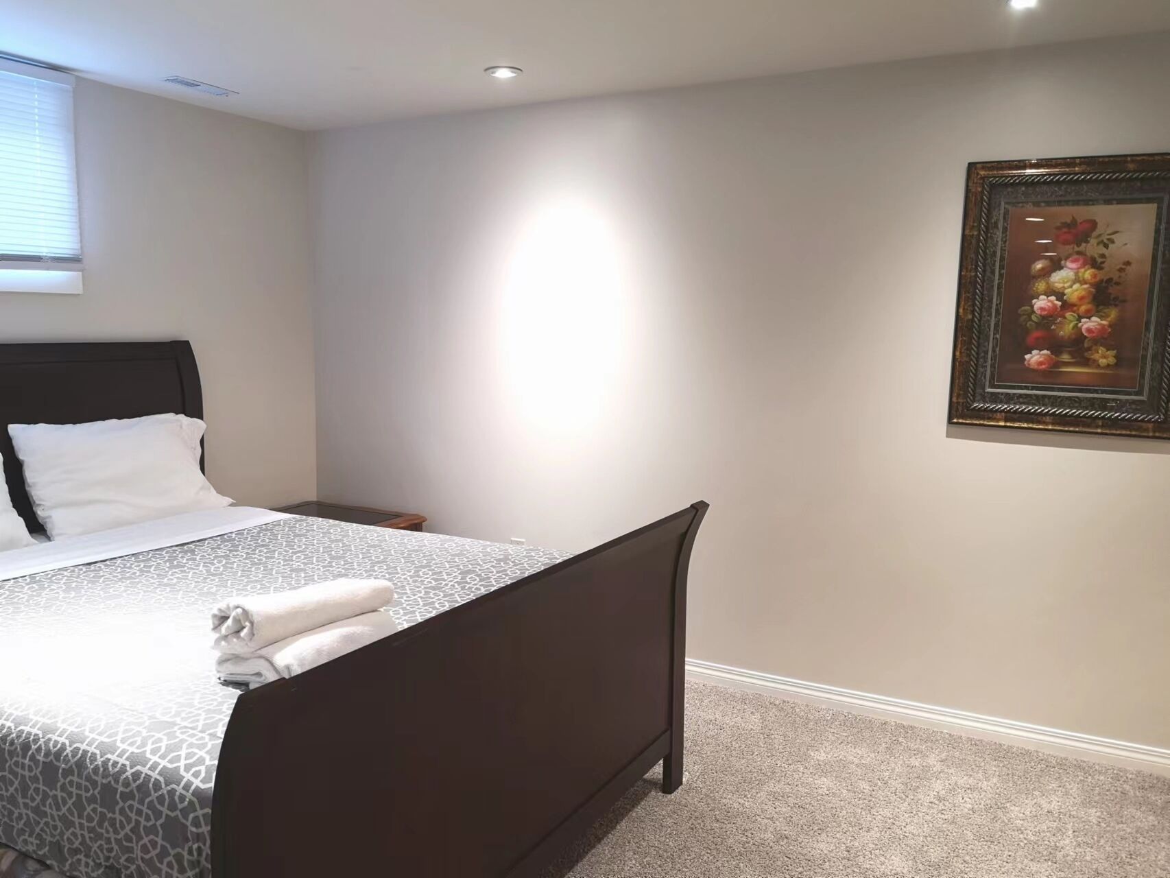 Una cama bien tendida con una colcha estampada y almohadas blancas, un cabecero oscuro, una mesita de noche pequeña y un cuadro con flores en la pared.