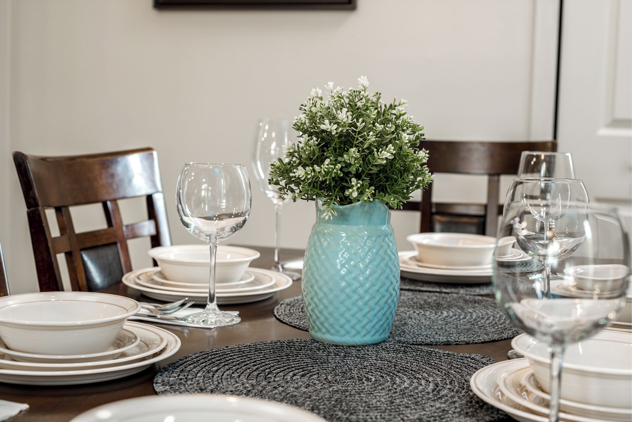 A dining table set with plates, bowls, and wine glasses, a blue vase with flowers, and a woven placemat.