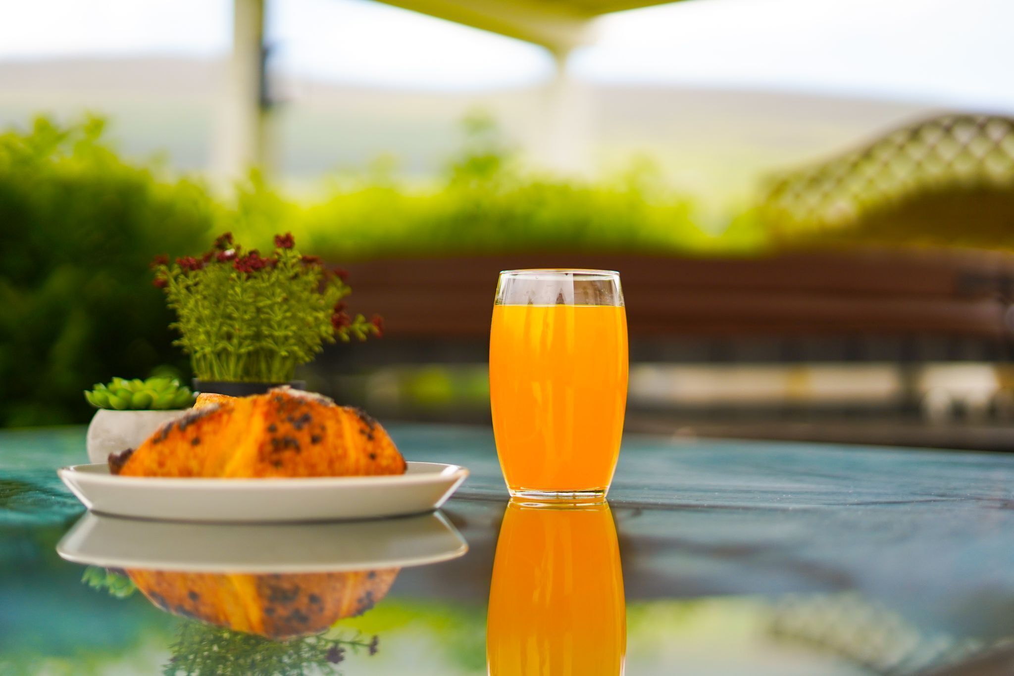 Un vaso de jugo de naranja sobre una superficie reflectante, con un plato de comida y una planta pequeña al fondo.