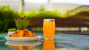 Un vaso de jugo de naranja sobre una superficie reflectante, con un plato de comida y una planta pequeña al fondo.