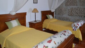 Une chambre avec deux lits en bois, chacun recouvert d’une couverture jaune et agrémenté d’un coussin à motifs floraux. Une table de chevet avec une lampe se trouve entre les deux lits.