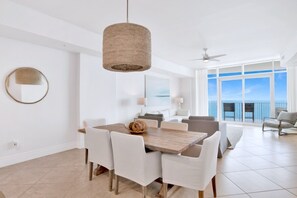 Un espace repas moderne avec une table en bois et des chaises blanches, un luminaire suspendu en fibres tressées, et une vue sur l’océan grâce à de grandes fenêtres.