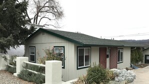 A small, single-story house with a green roof and white siding. It features a front porch with a white railing and a small garden area with various plants and shrubs. - Cozy 1 Bedrm/1 Bth, Walk to Downtown, Pet Friendly, Fenced yard, Covered Patio (Prescott)