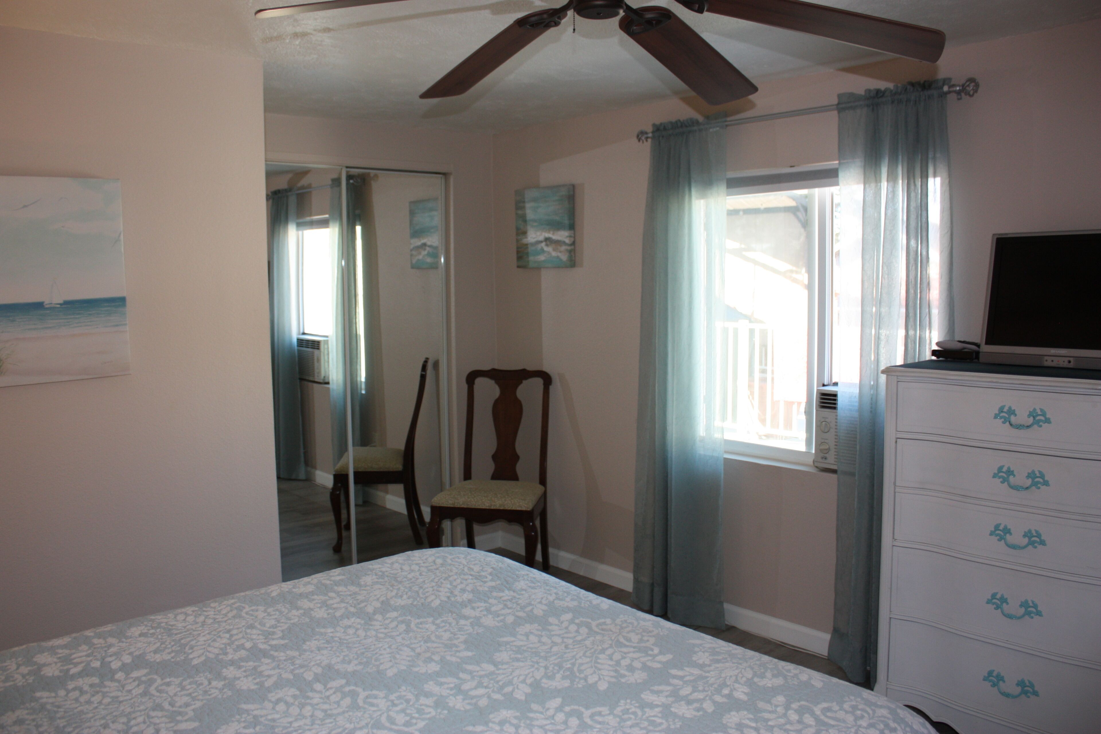 Un dormitorio con cama, cómoda, silla, ventilador de techo y ventana con cortinas.