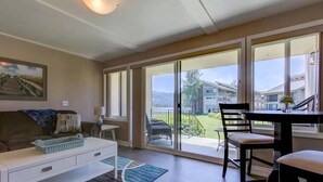 A living room with a sofa, a coffee table, and a dining area with chairs and a table. There is a large sliding glass door leading to a balcony with a view of a building and a garden. - Downtown Sandpoint Lakeside Condo (Sandpoint)