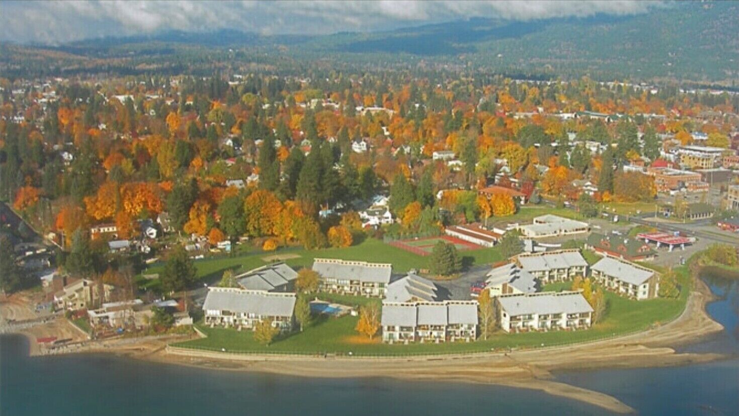 An aerial view of a coastal town with a marina, surrounded by autumn-colored trees and residential buildings.