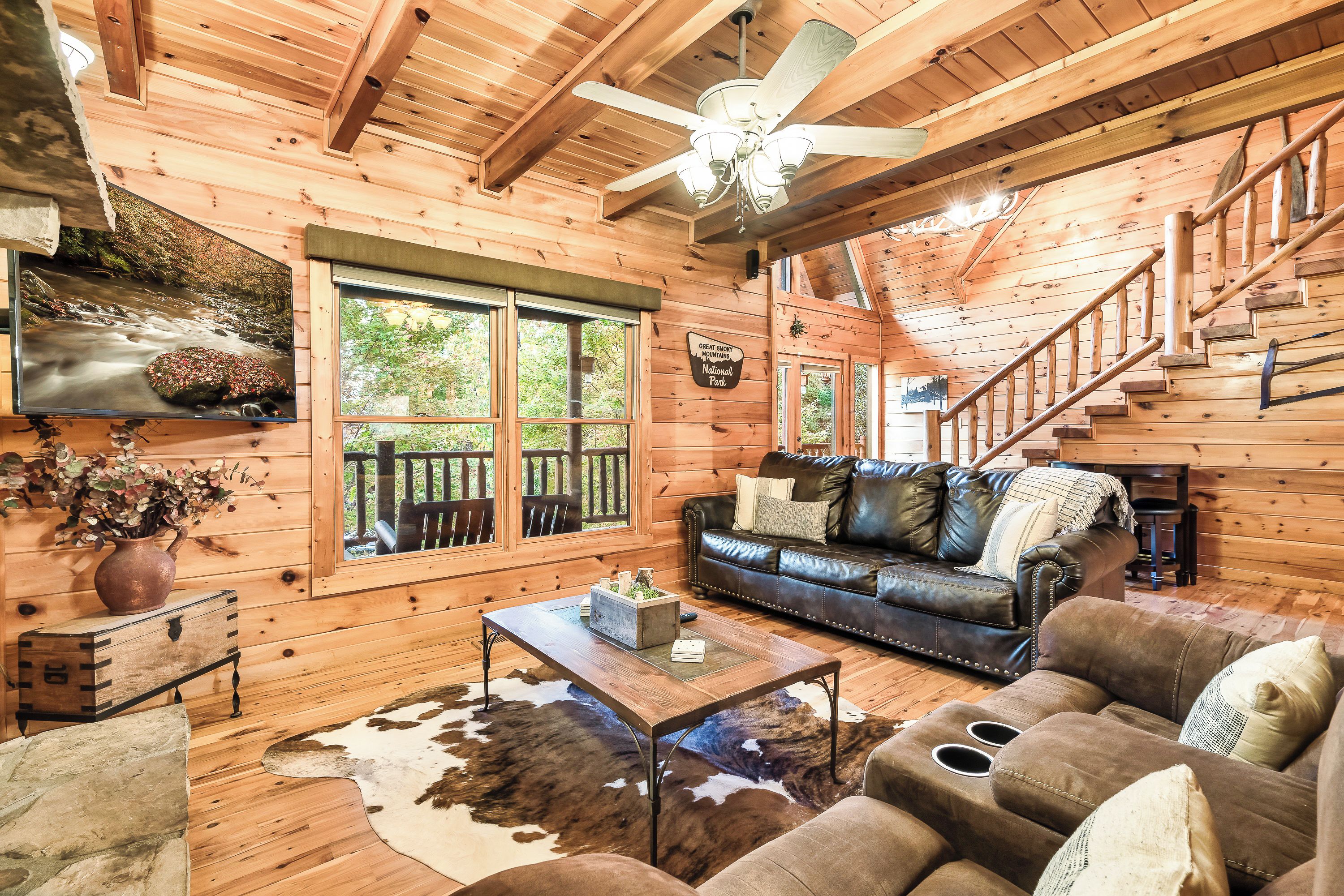 L’intérieur d’une cabane en rondins, avec un plafond en bois, un ventilateur de plafond, une cheminée, un canapé en cuir, une table basse et une grande fenêtre.