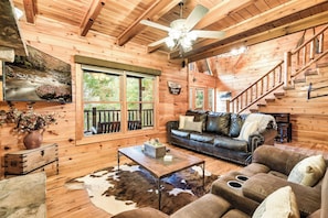 A log cabin interior with a wooden ceiling, a ceiling fan, a fireplace, a leather sofa, a coffee table, and a large window.