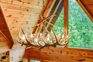 A chandelier made of antlers hanging in a wooden room with a window overlooking greenery.