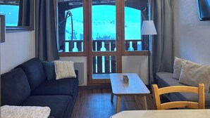 A cozy room with a wooden ceiling, a sofa, a table, and a chair. There are curtains and a window with a view of a snowy landscape.