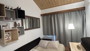 A cozy room with a bed, a small table, and a TV. There are shelves with decorations and a window with curtains.