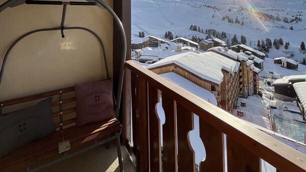 A wooden balcony with a ski lift chair, a view of a snowy mountain town, and a clear sky.