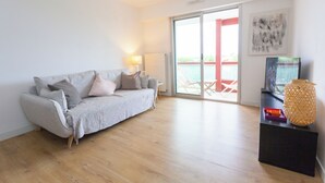A modern living room with a grey sofa, a small side table, and a large window with a view of the outdoors. - Appartement 2 Chambres Séparées Denviron 60m2 Anglet-plages (Anglet)