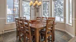 A dining area with a wooden table and chairs, large windows with a snowy view, and a chandelier. - Desirable Knoll Home with Hot Tub, BBQ, Laundry - Open Den with Sofa Bed (Silver Star Mountain)