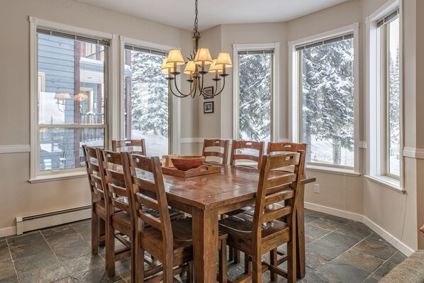 Une salle à manger avec une table et des chaises en bois, de grandes fenêtres donnant sur des arbres couverts de neige, et un lustre.