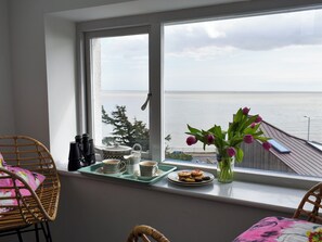 Un balcon avec vue sur la mer, une chaise en osier, une table sur laquelle se trouve un plateau avec des tasses et des pâtisseries, et un vase rempli de fleurs.