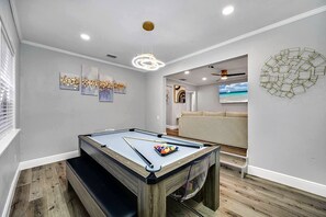 Una mesa de billar en una habitación con ventilador de techo, obras de arte en las paredes y vista a una playa a través de una ventana.