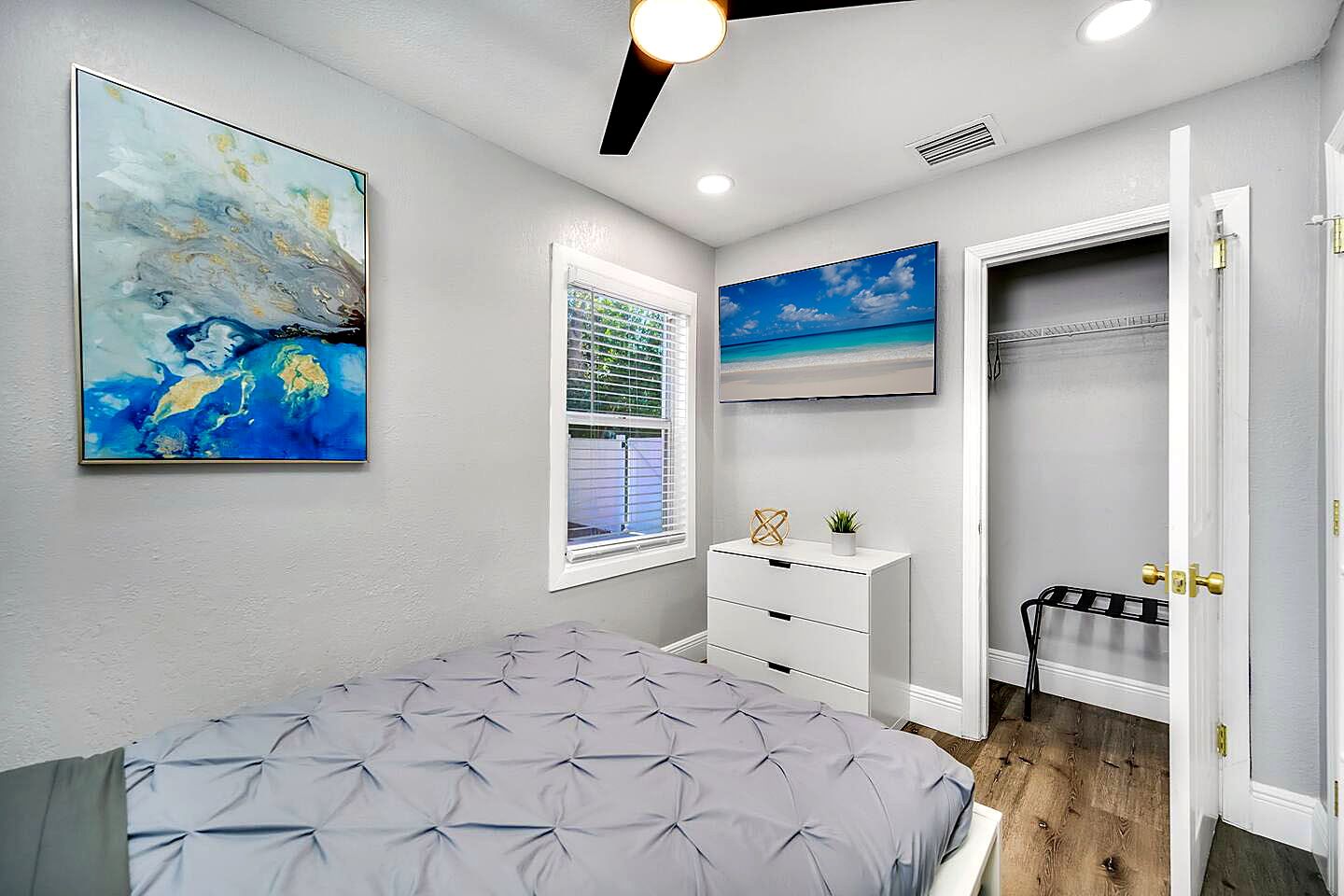 Un dormitorio con cama, ventana con persianas, televisor montado en la pared, armario con puerta y ventilador de techo.
