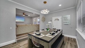 Un comedor moderno con una larga mesa de madera dispuesta para seis comensales, un banco y vista a la playa a través de un ventanal abierto.