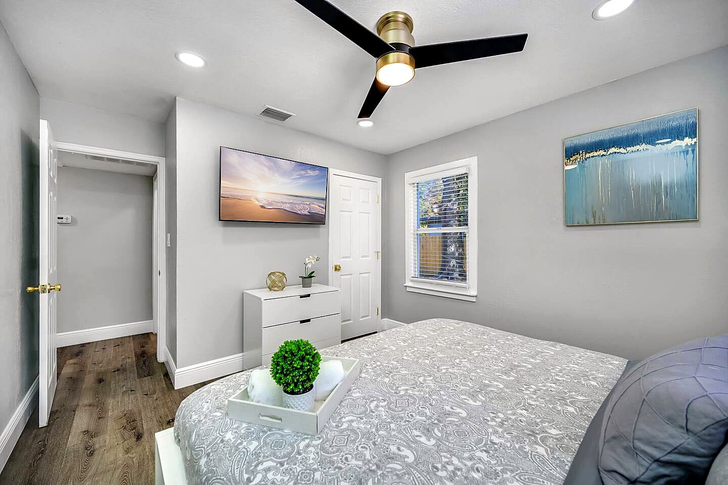 Une chambre à coucher avec un lit, un ventilateur de plafond, une fenêtre avec des stores, un tableau et une porte.