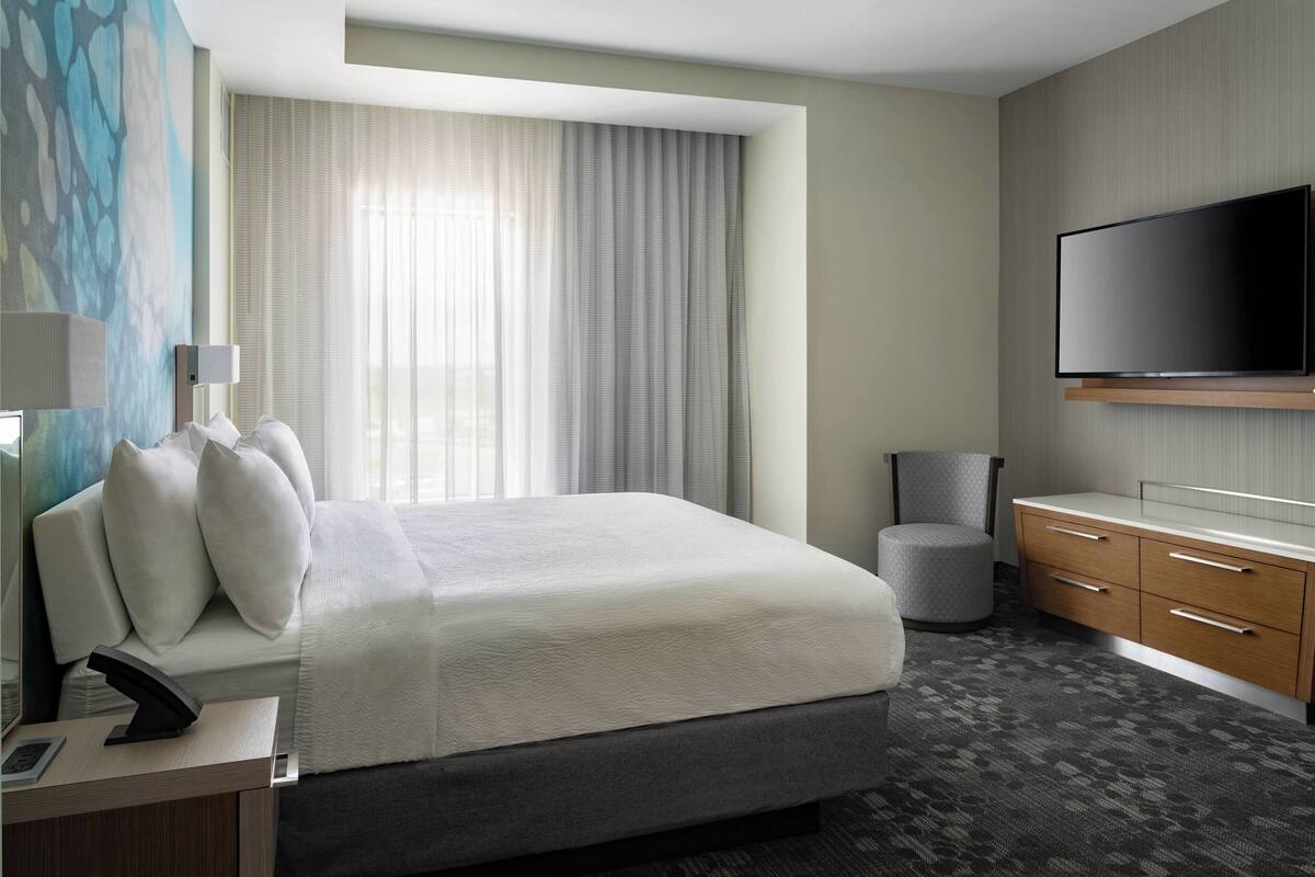 Habitación de hotel moderna con una cama grande, un televisor de pantalla plana montado en la pared, un escritorio con lámpara y vistas al exterior a través de cortinas traslúcidas.