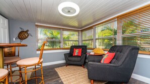 Ein gemütliches Zimmer mit Holzboden, Deckenventilator und großen Fenstern mit Jalousien. Zwei Stühle, ein kleiner Tisch und ein dekoratives Wandbild sind vorhanden.